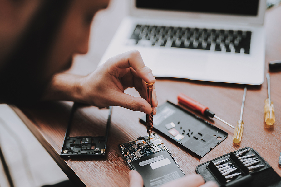 A technician uses a precision screwdriver to repair the internal components of a smartphone on a wooden desk, surrounded by small tools, spare parts, and an open laptop.
