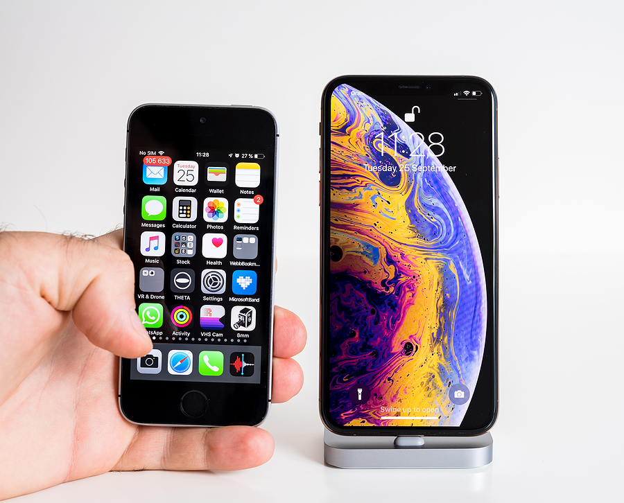 Hand holding a small older iPhone next to a larger modern iPhone on a stand, showing the size and design differences between an old iPhone and a newer model.